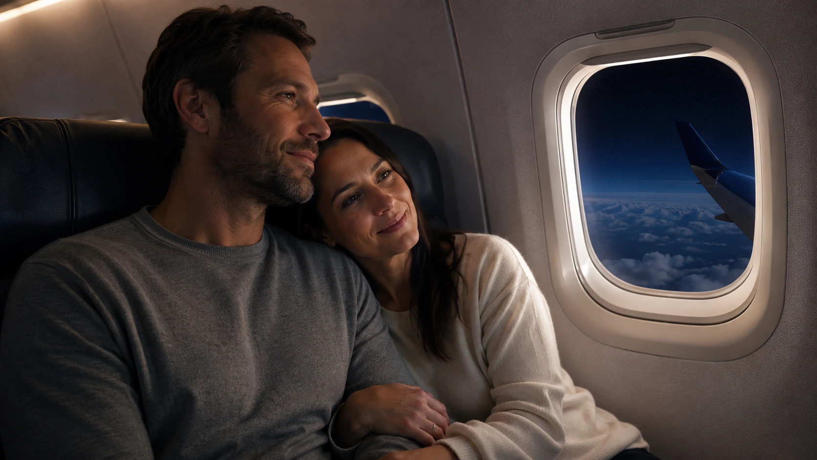 couple cuddling in airplane seats beside window at night, looking peaceful as wing and clouds appear outside
