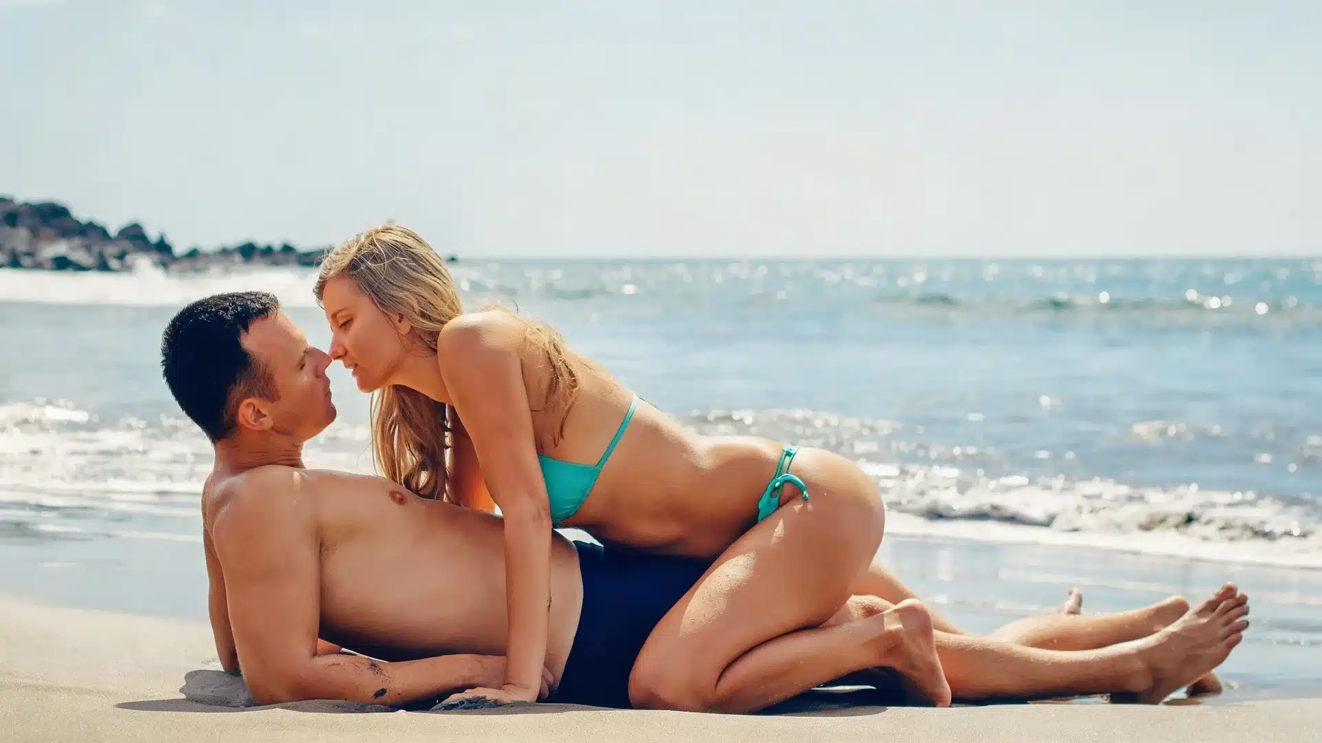 couple in swimwear lies closely on a sunny beach near ocean waves, sharing an intimate moment by the shoreline together