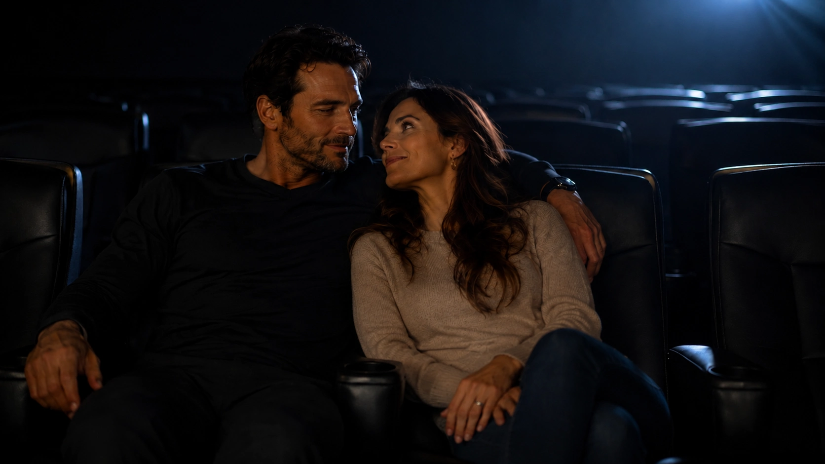 couple shares a quiet moment in a movie theater, smiling at each other while seated comfortably under soft screen light