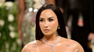 Demi Lovato wearing a sparkling diamond necklace and earrings, with tattoos visible on her shoulder and chest.