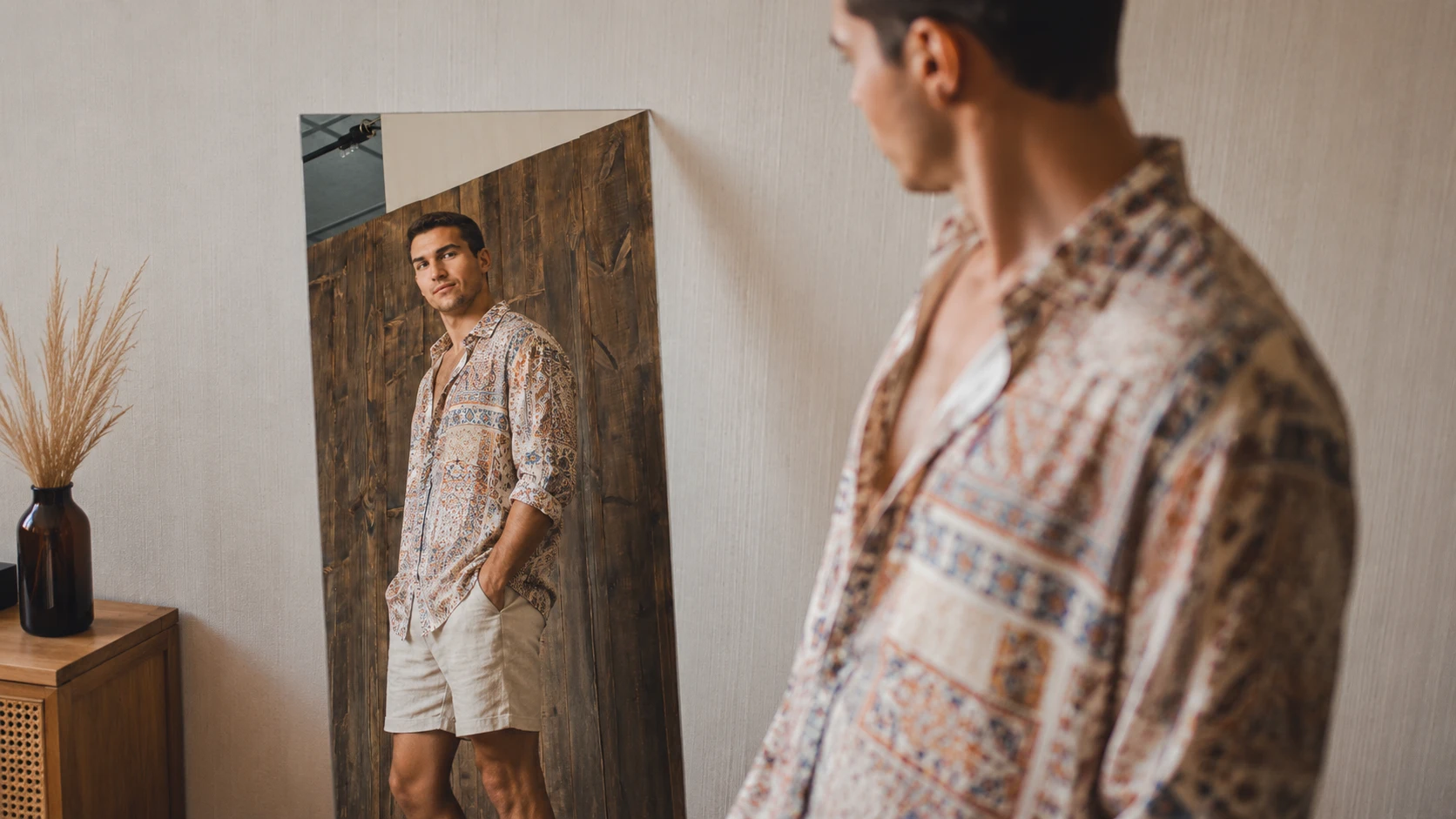 egoistic man in bohemian shirt and shorts looks at himself in a mirror inside a warm modern room