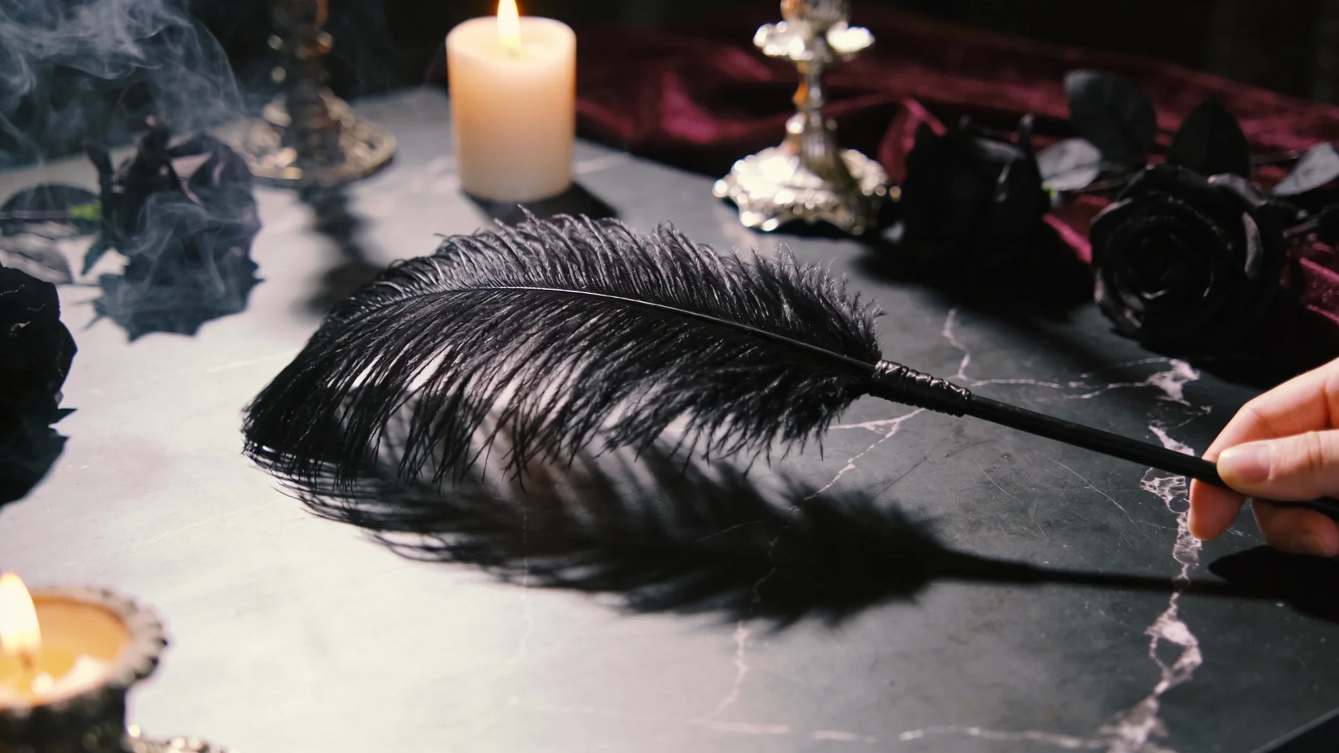 hand holding a black feather quill over dark marble, surrounded by lit candles, smoke, black roses, and velvet decor
