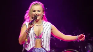 JoJo Siwa sings into a microphone on stage wearing a sequined vest and matching sparkly outfit with bright pink lighting.