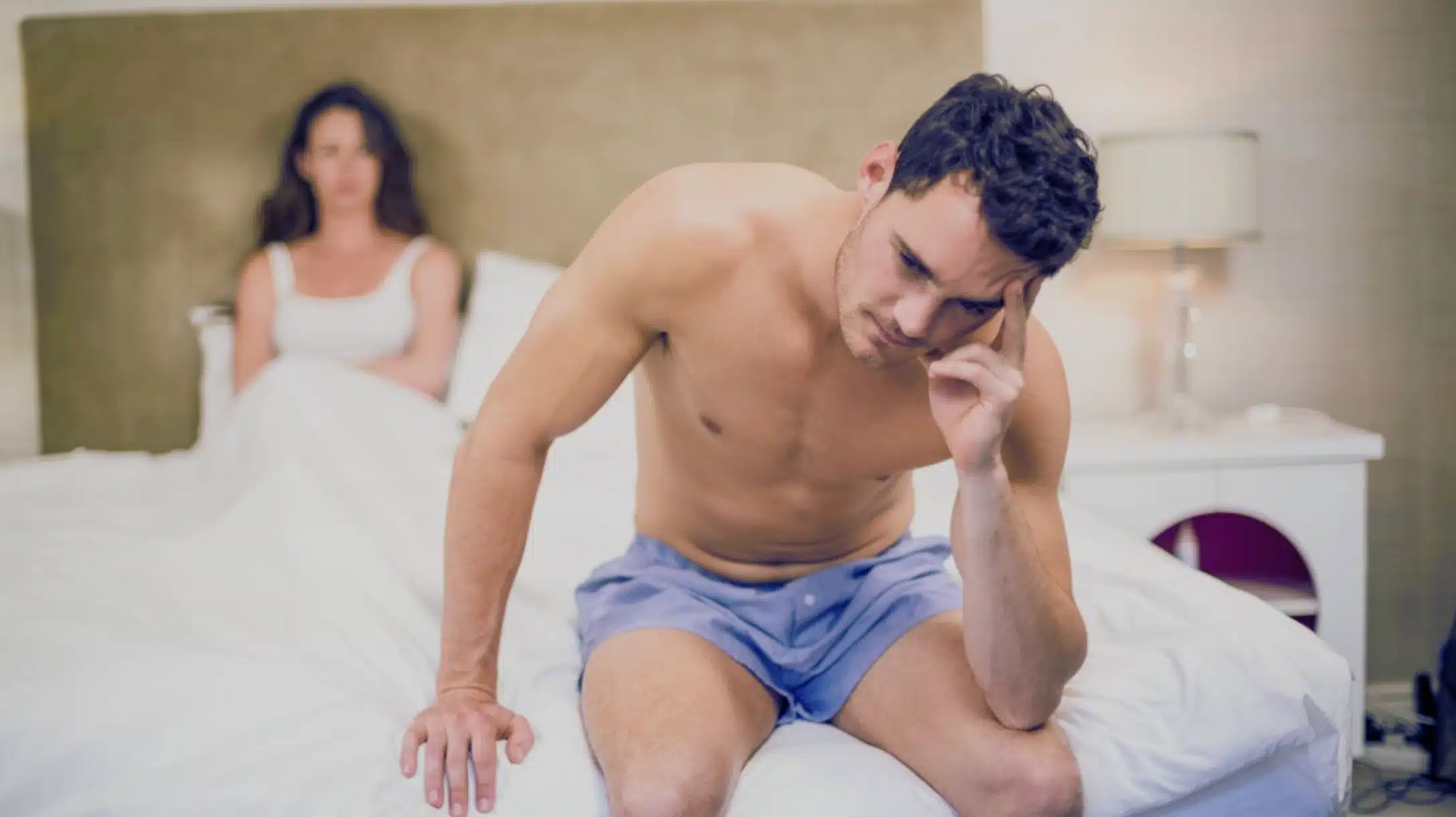 man sitting on bed looking troubled after intimacy while woman sits behind him, suggesting post nut clarity