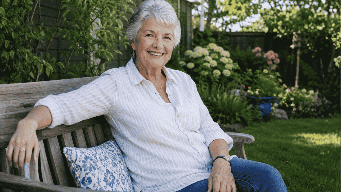 older woman smiling on a backyard bench in a sunny garden with flowers