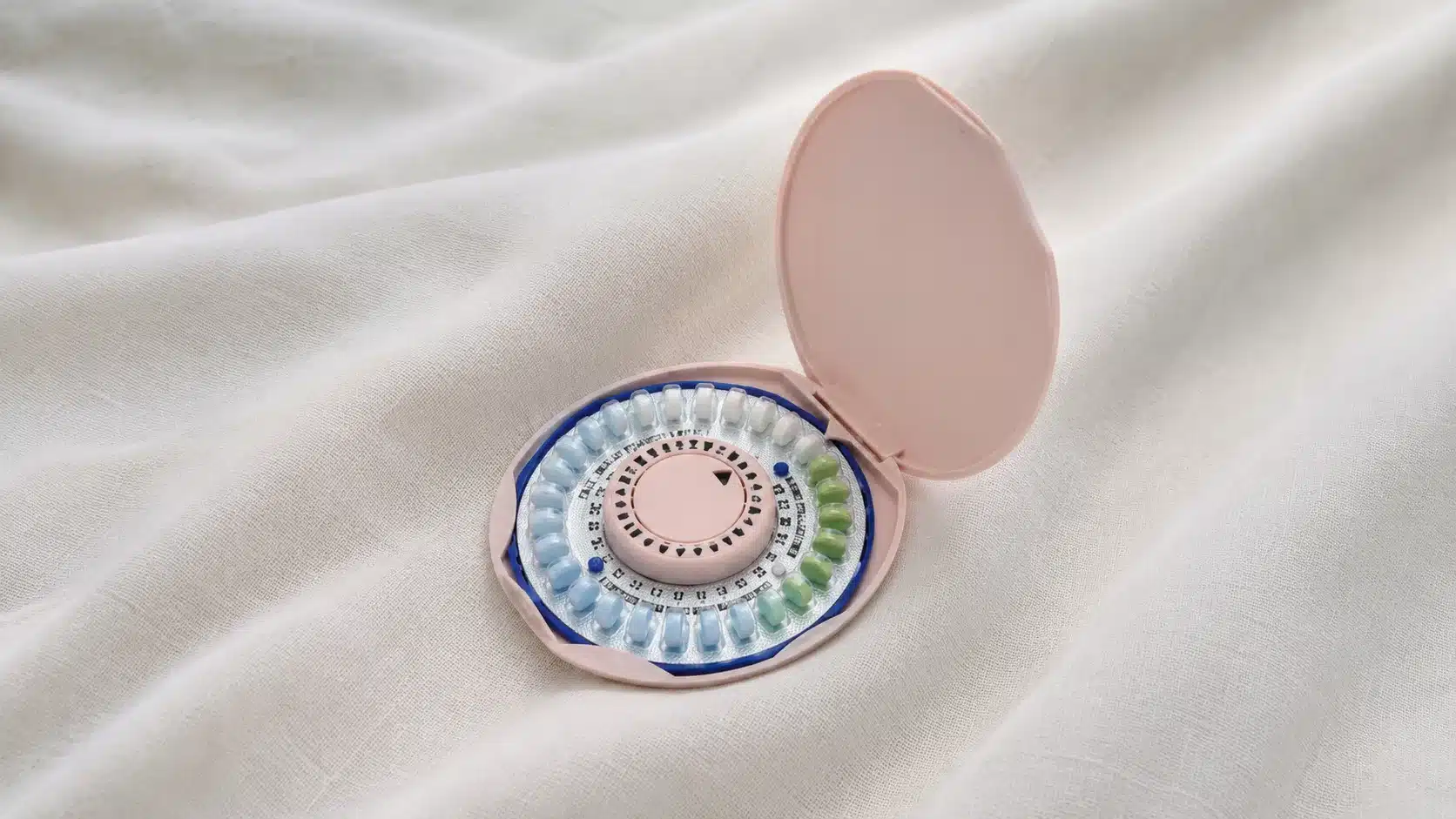 open pink birth control pill case with colorful circular tablets resting on white fabric, shown in realistic close-up