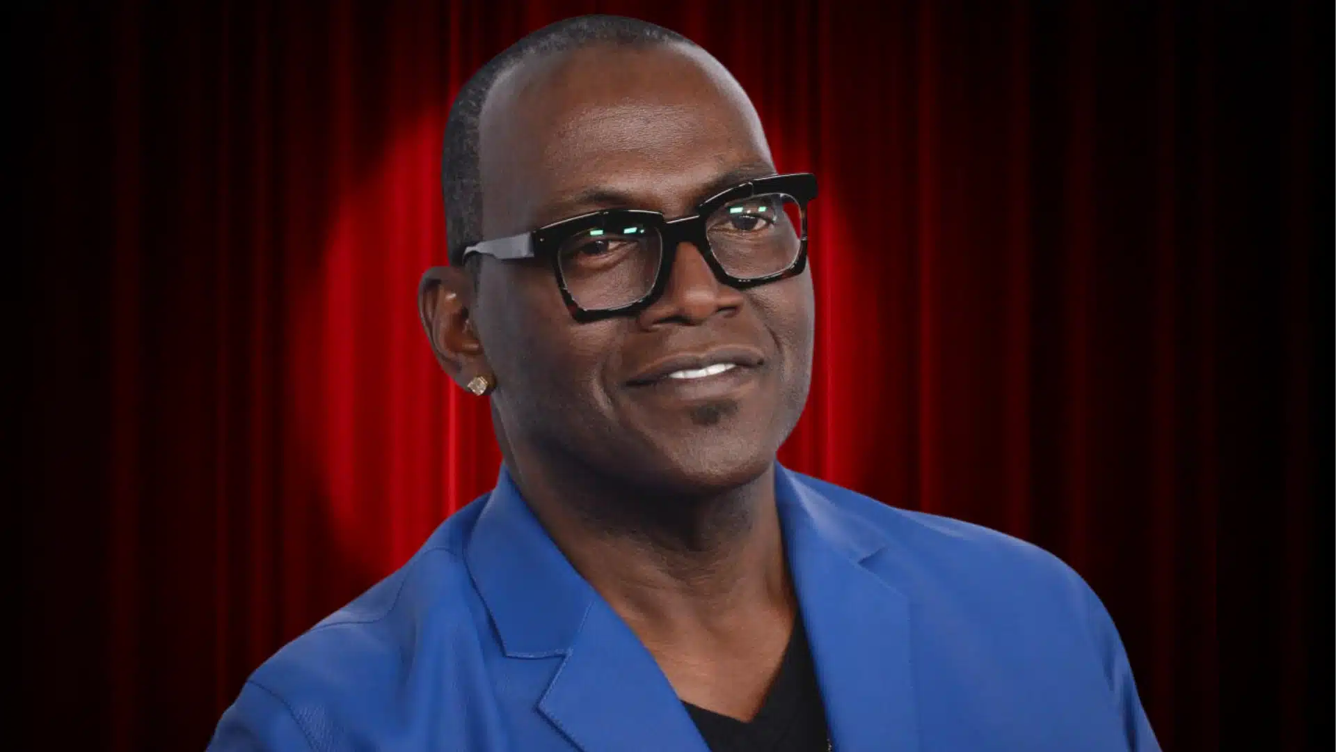 Randy Jackson in black glasses, a blue blazer, and an earring, posing against a red curtain background