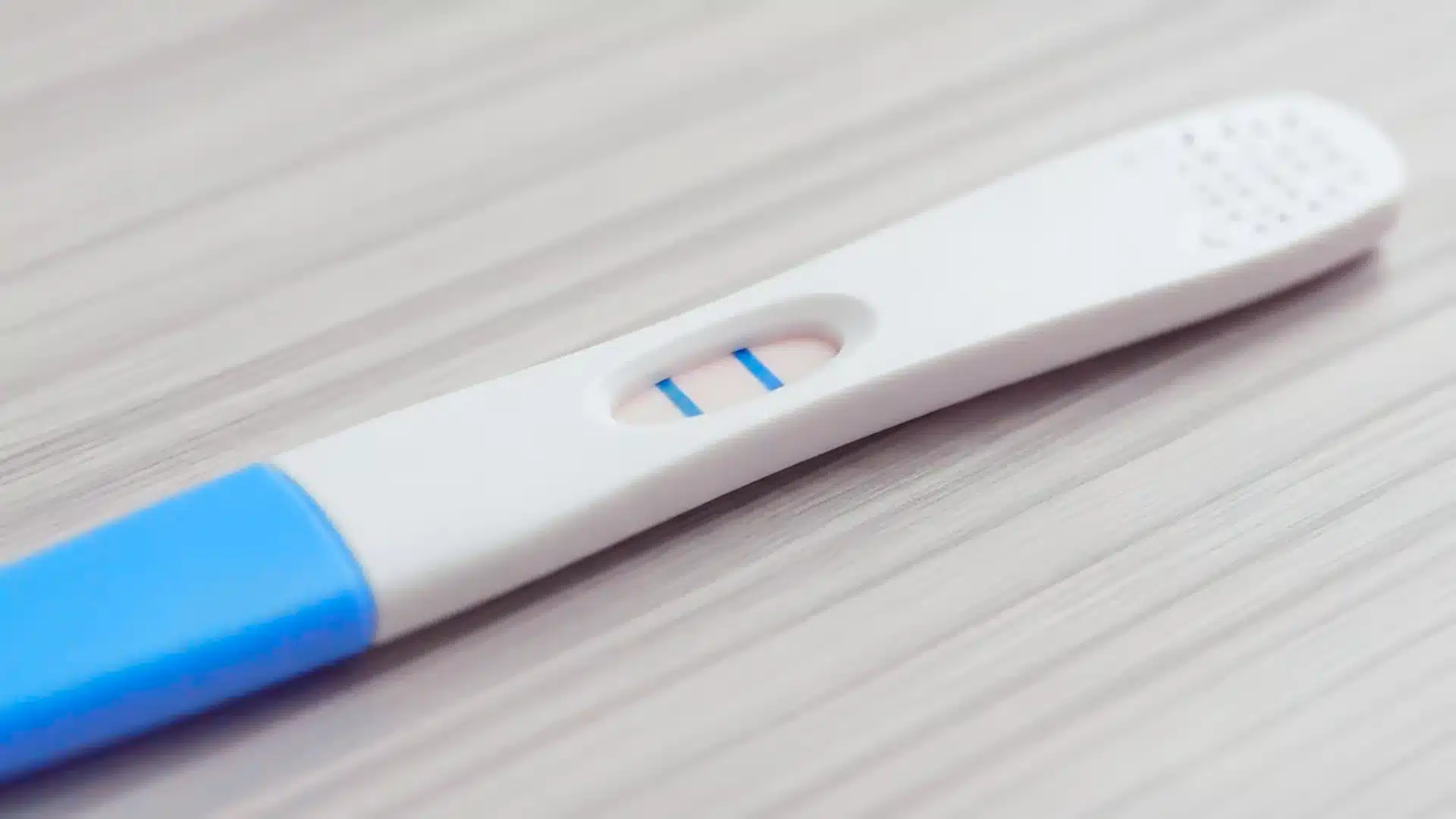 A pregnancy test shows two blue lines, indicating a positive result. The test, with a blue and white handle, rests on a light wood surface.