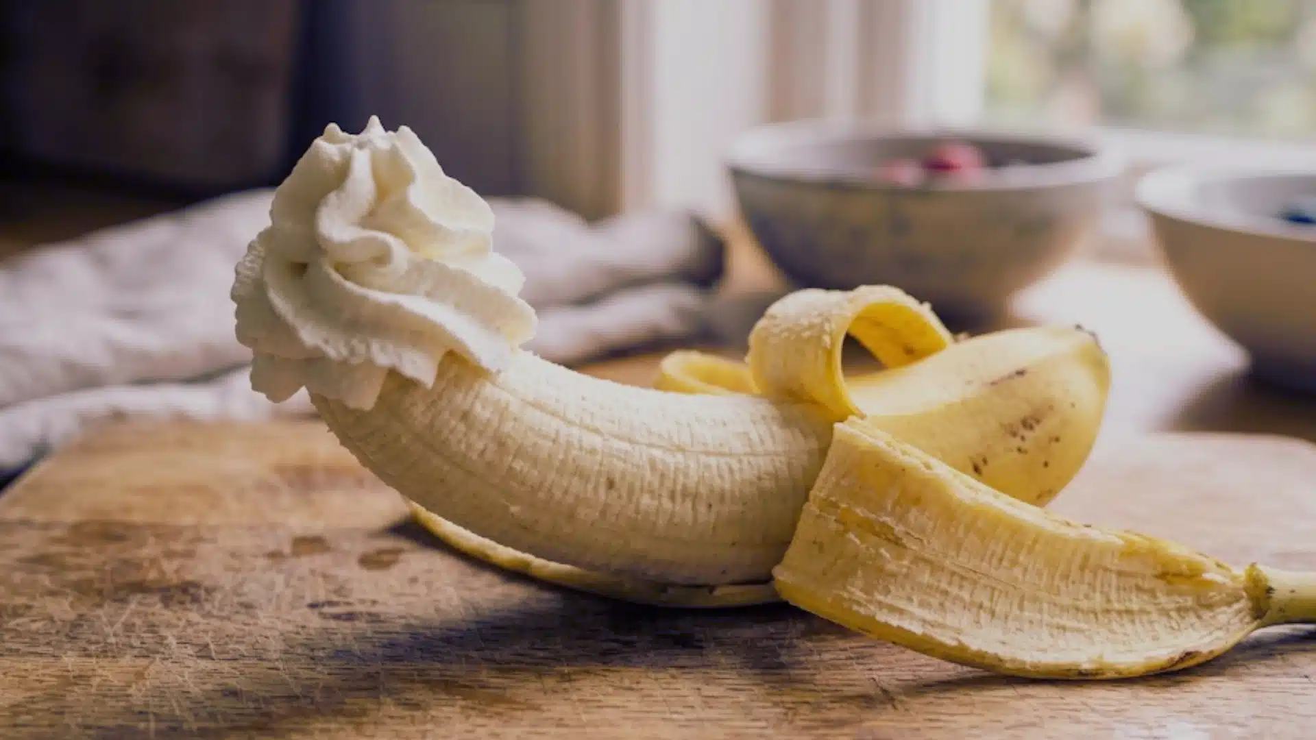 A peeled banana topped with whipped cream on a wooden surface, with bowls blurred in the background.