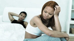 woman sits looking withdrawn while man watches from bed, suggesting post nut clarity quiet relationship discomfort