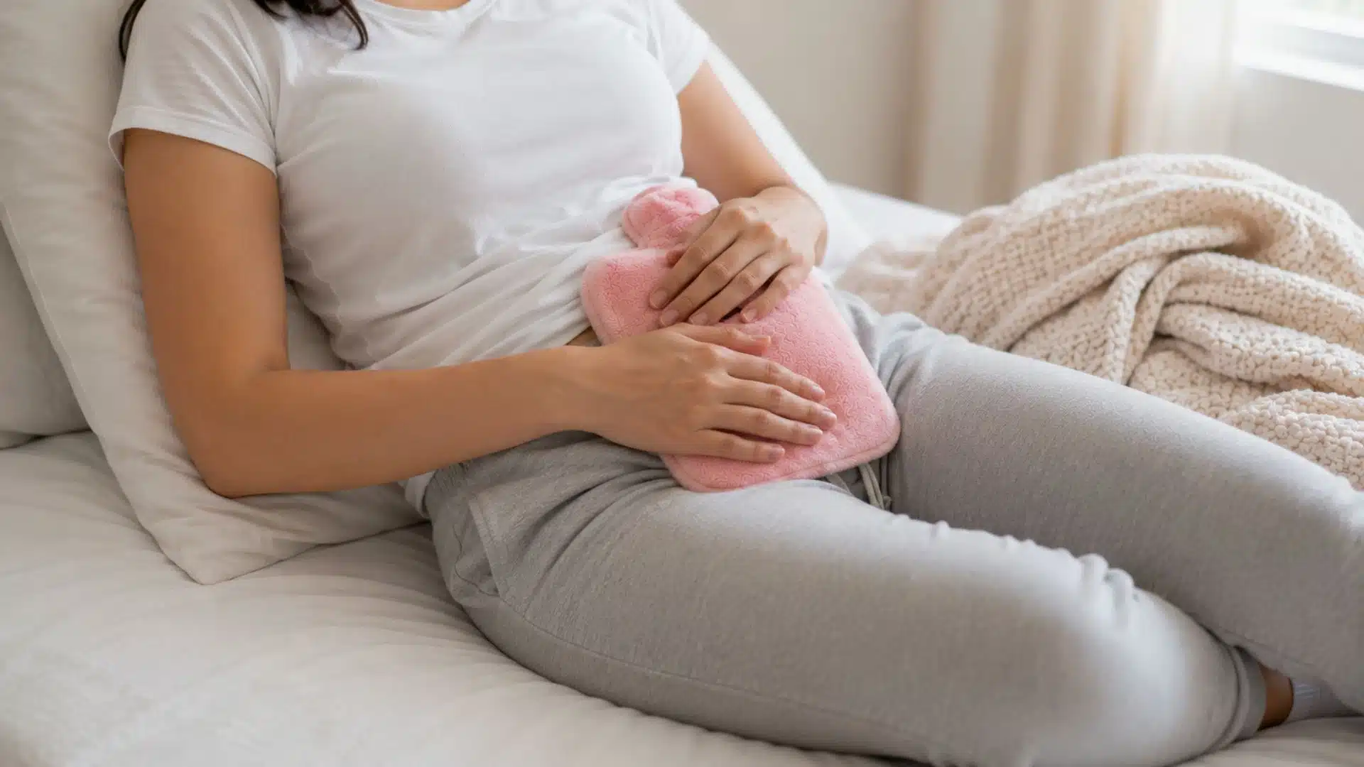 A woman sits on a bed, holding a pink hot water bottle, looking relaxed and comfortable.
